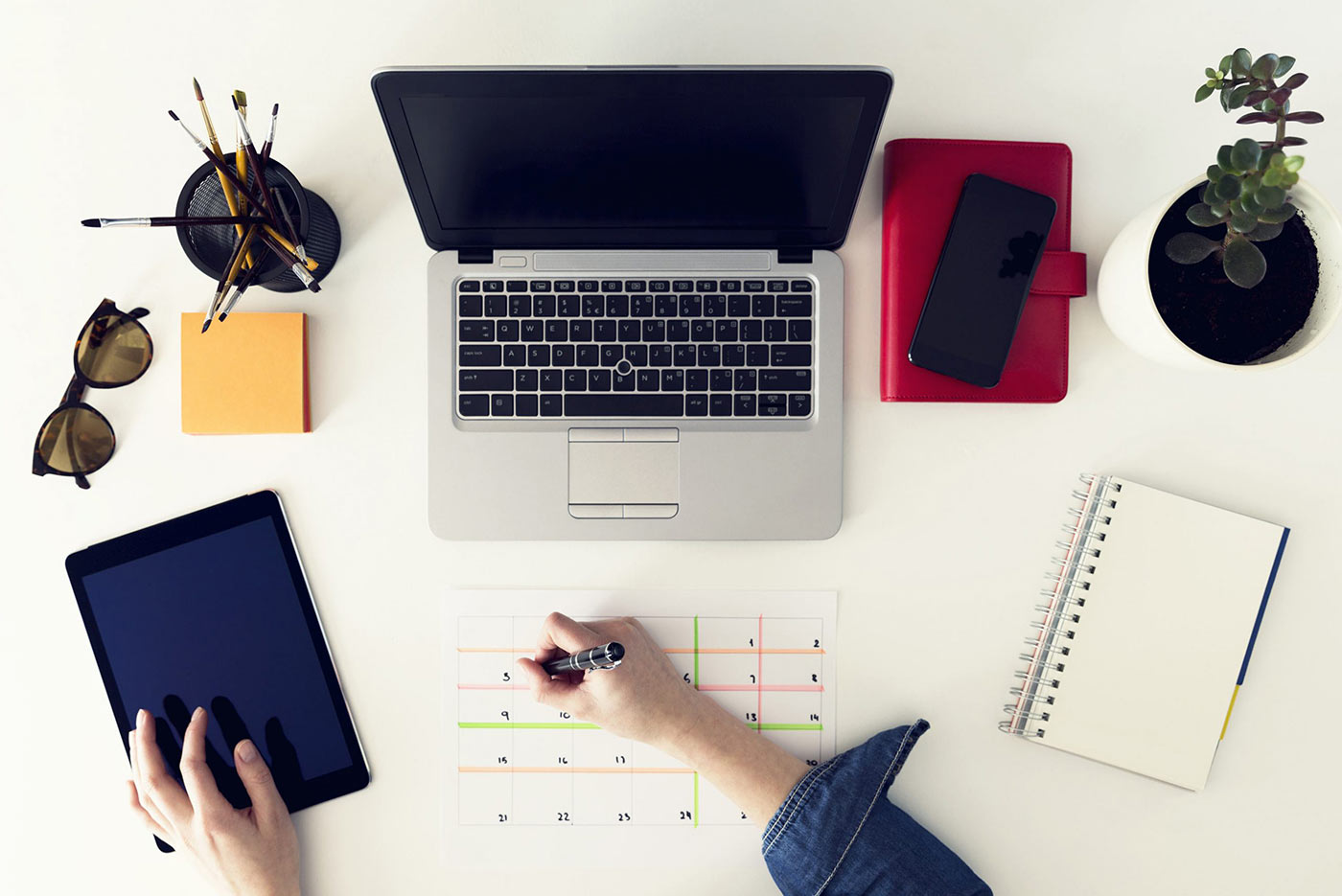 Desk layout with laptop, stationery, mobile, plant and man drawing in calendar